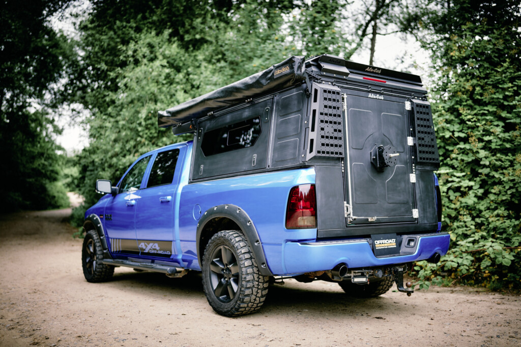 Blauer Dodge RAM 1500 mit Alu-Cab ModCAP Aufbau, Heckansicht mit geöffneter Seitenklappe auf einem Waldweg
