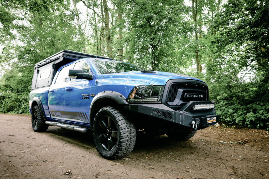 Blauer Dodge RAM 1500 mit Alu-Cab ModCAP Camper-Aufbau, Frontansicht im Wald